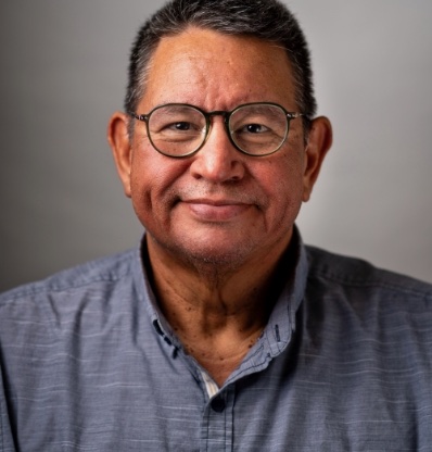 An elderly man with glasses and a grey shirt is smiling slightly against a plain background.
