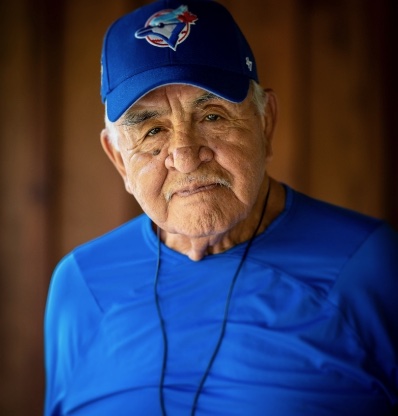 An elderly man wearing a blue cap and shirt stands against brown backdrop with his hands clasped together.