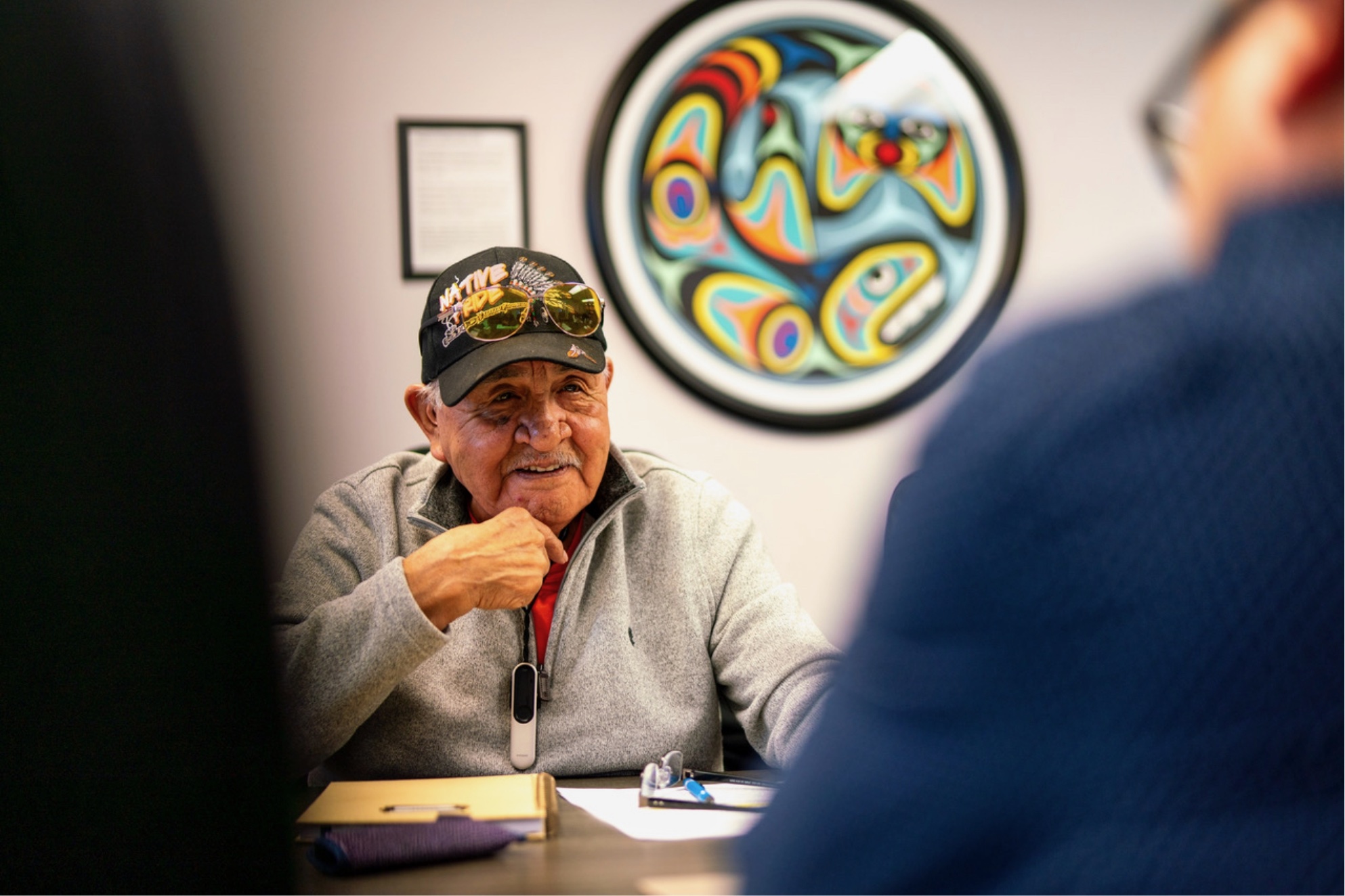 An elderly man wearing a cap is sitting at a table with papers and art behind him.