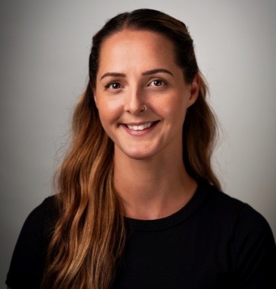 A person with long hair and a smile is standing against a plain background.
