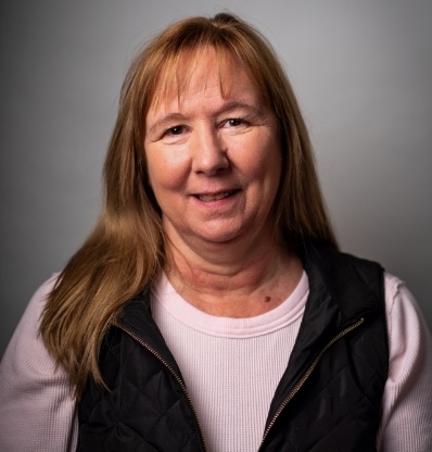 A person with long hair and a vest is smiling against a plain background.