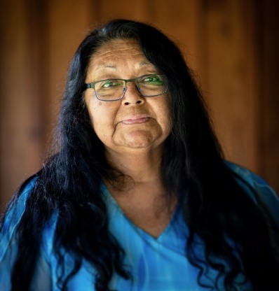 A person with long hair wearing glasses and a blue shirt stands against a brown background.