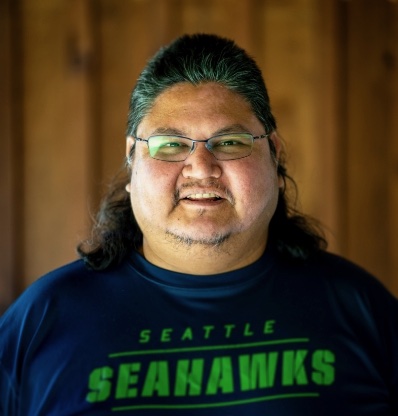 A person with long hair wearing a Seahawks shirt is standing against a brown background.