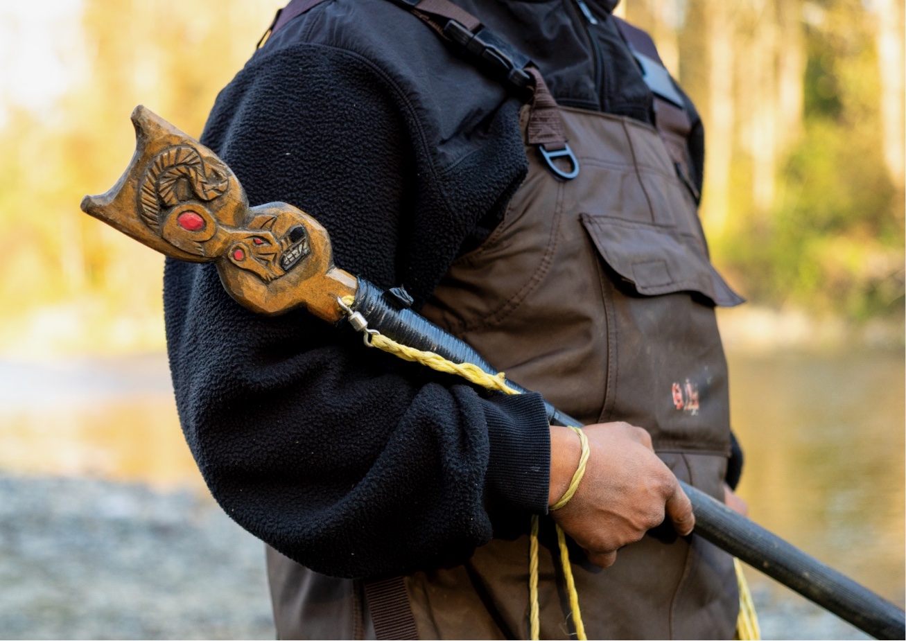 A person is holding a staff with an animal head at the top while wearing brown overalls and a black jacket.