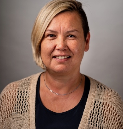 A person with a necklace and cardigan is smiling against a grey background.