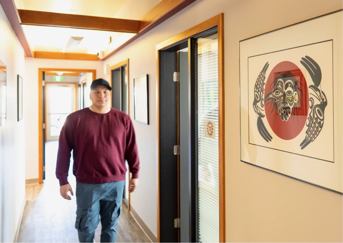 Man walking down hallway with framed artwork and door.