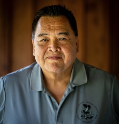 Man wearing a shirt with logo and standing against brown background.