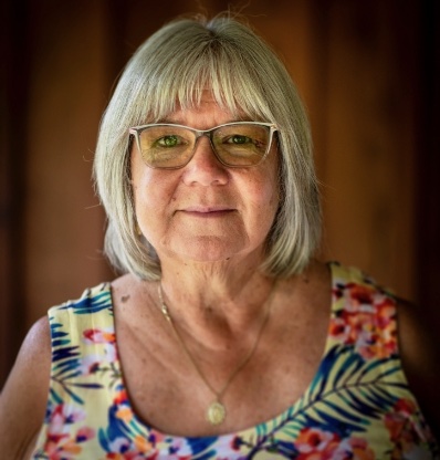 An elderly woman with glasses and a floral dress is smiling at the camera.