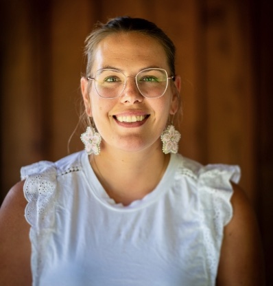 Woman with glasses and earrings smiling against brown background.