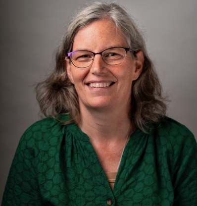 A woman with glasses and a green shirt is smiling against a grey background.
