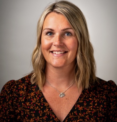 A woman with blonde hair and a necklace is smiling against a plain background.