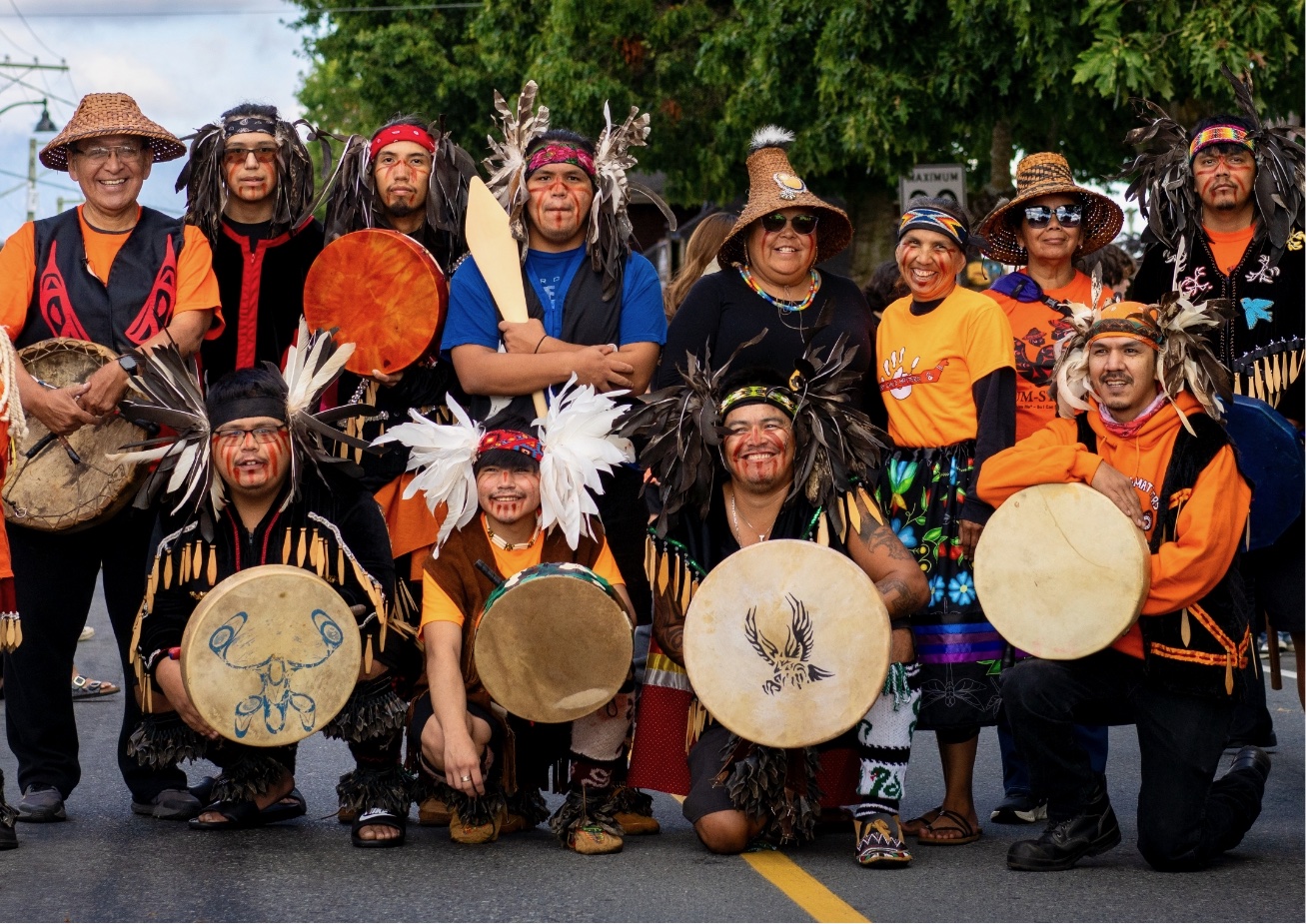 A group of people wearing traditional clothing and holding drums pose for a picture with trees in the background.