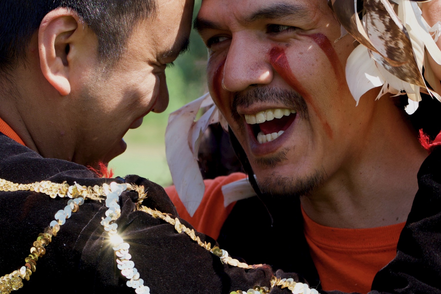 Two men with red marks on their faces are laughing and interacting closely outdoors.