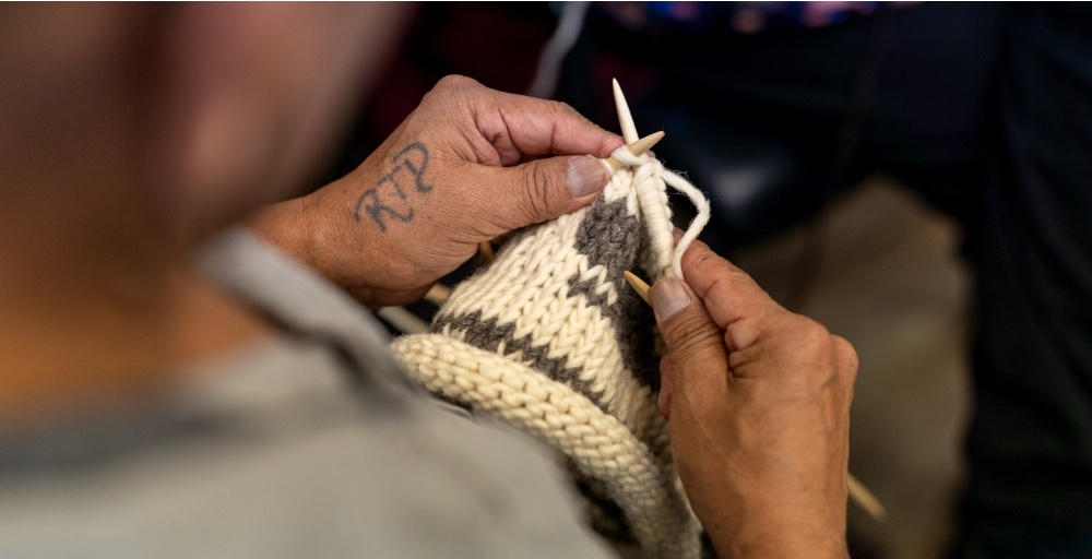 A person is knitting with white and brown yarn while wearing a tattooed hand.