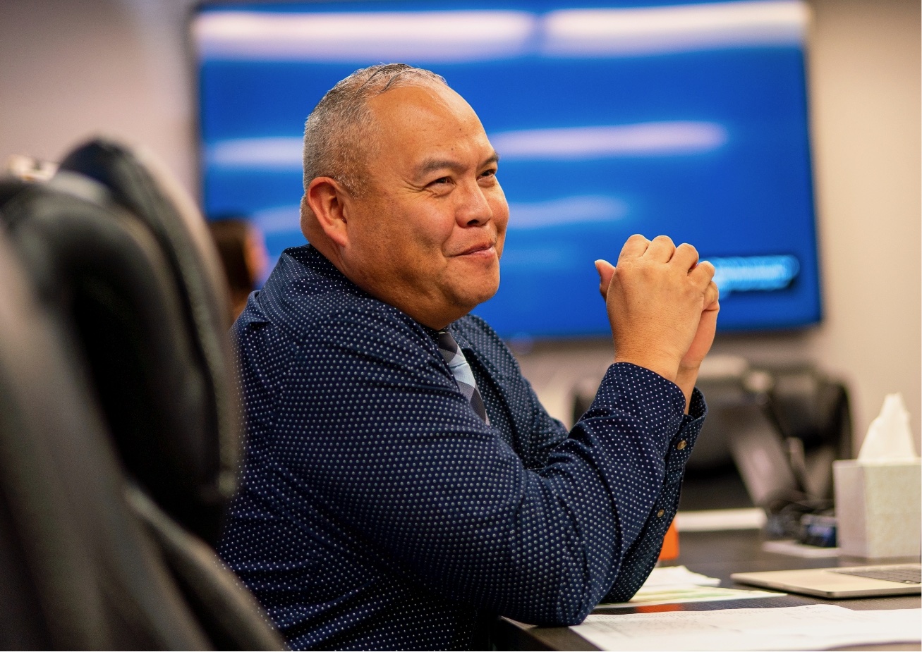 Man sitting at table with tv behind him and white wall.