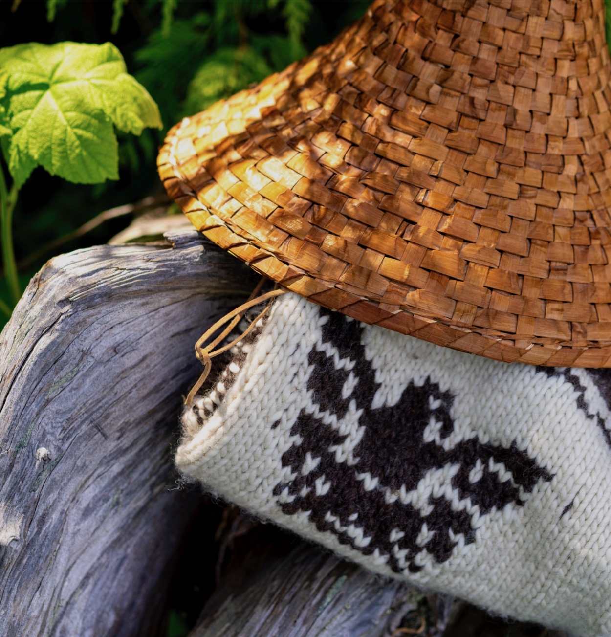 A woven hat and a knitted scarf are placed next to each other outdoors with green foliage in the background.