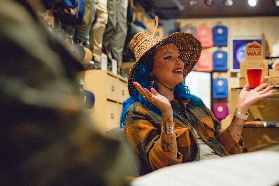 A person with blue hair and a straw hat is holding up an object while sitting at a table.