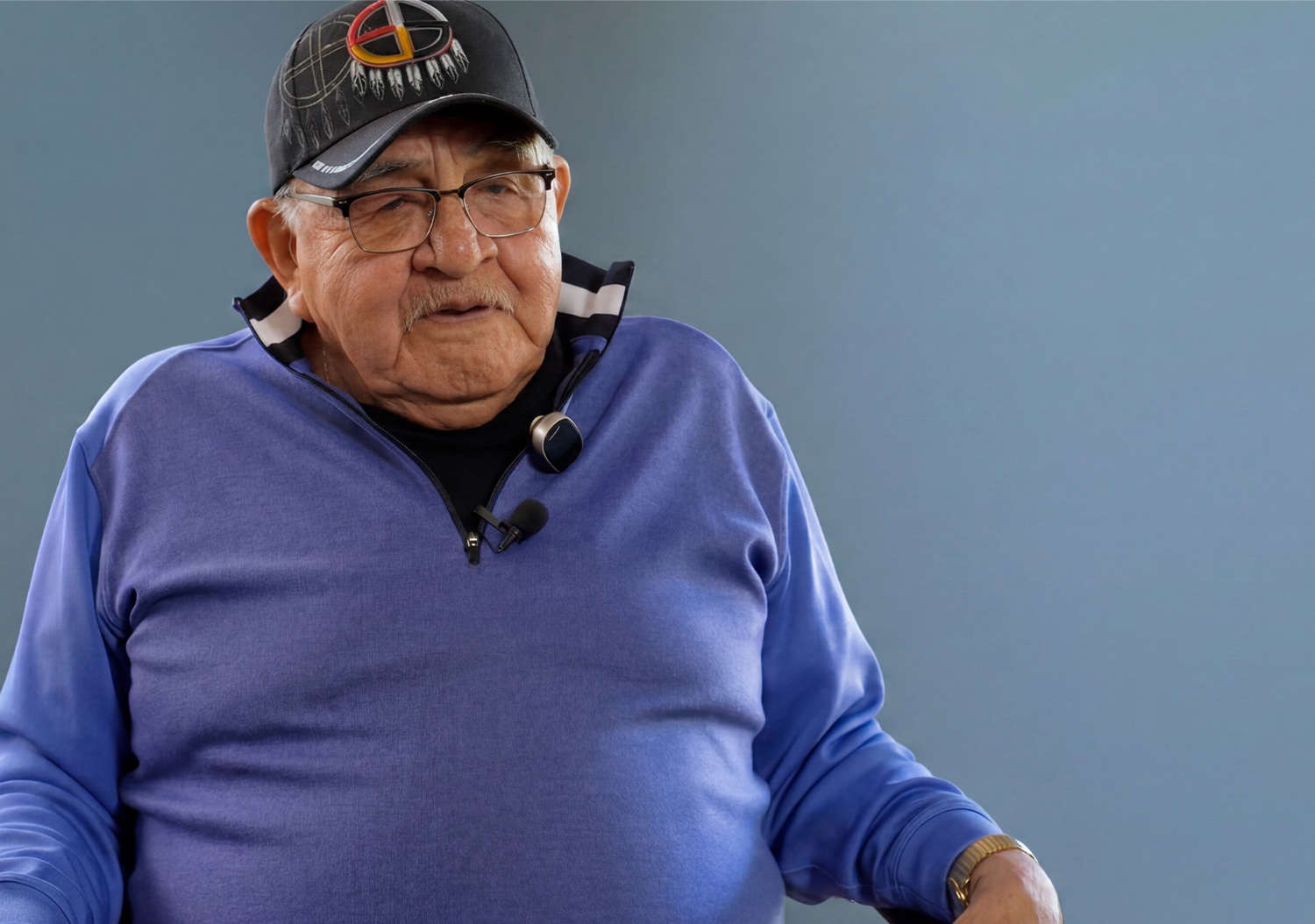 An elderly man wearing a cap and sweater is standing against a light blue background with his hands raised slightly above him.